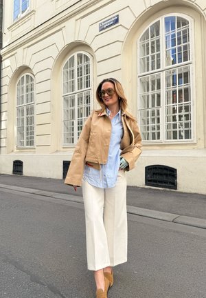 Chaqueta corta color beige con un acento rojo, camisa de rayas azules, pantalones anchos blancos y zapatos de ante marrones. Las gafas de sol completan el conjunto.