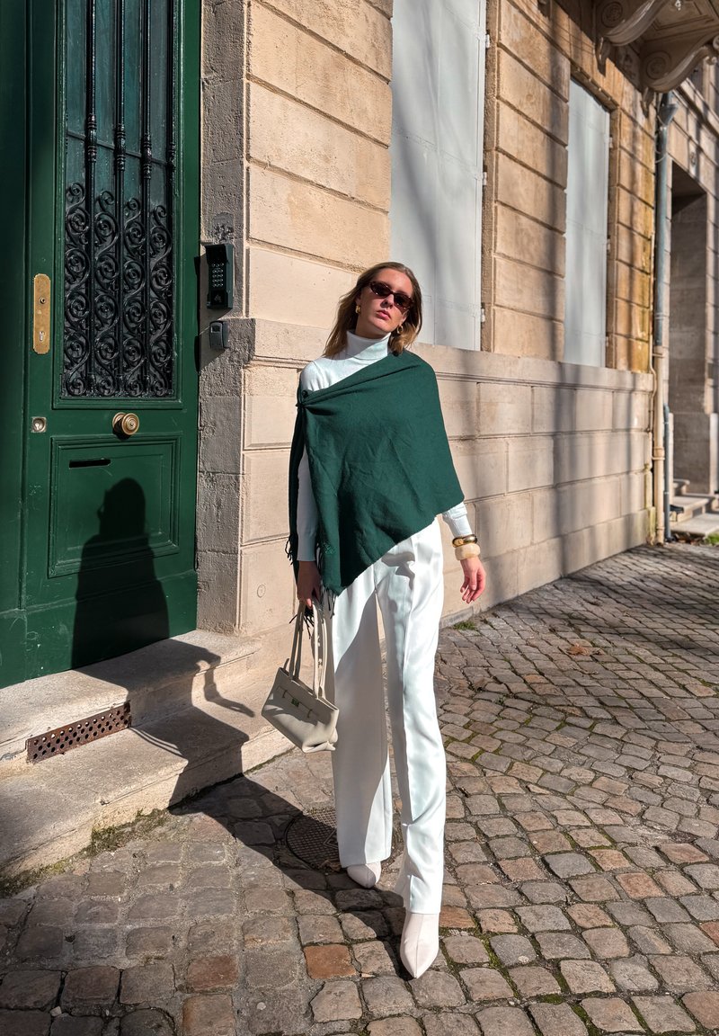 Femme portant un pantalon blanc, un col roulé blanc, un châle vert, des lunettes de soleil et des bottes beiges, marchant sur une rue pavée à côté d'une porte verte et d'un bâtiment en pierre beige.