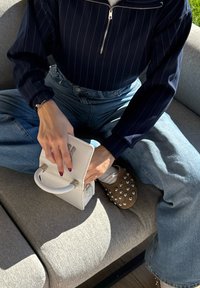 Osoba v tmavém pruhovaném topu a modrých džínách, sedící na pohovce s překříženýma nohama, držící bílou kabelku, s hnědými slip-on botami s cvočky.