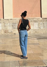 Top senza maniche nero, jeans a vita alta blu, scarpe slip-on a motivi bianco e nero, in piedi contro un muro di colore chiaro.