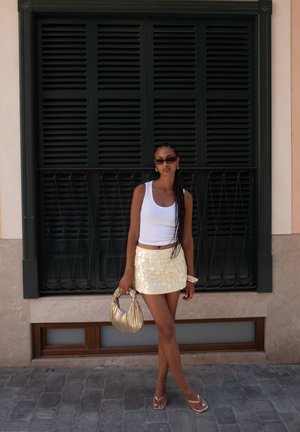 Top ajustado blanco, mini falda de lentejuelas doradas, bolso metálico dorado, gafas de sol negras, de pie contra un fondo de persianas verdes.
