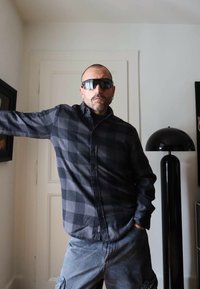 Camicia di flanella a quadri neri e grigi con bottoni, indossata con pantaloni cargo grigi. Occhiali da sole scuri; sullo sfondo una lampada nera e un'opera d'arte murale.