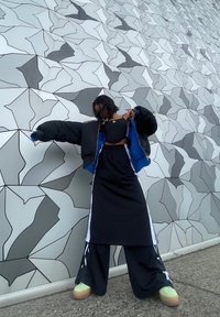 Doudoune noire avec doublure bleue, haut court noir et jupe longue noire avec des rayures latérales. Le mannequin porte des chaussures à plateforme vertes. Fond de mur abstrait.