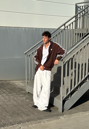 Chaqueta deportiva marrón con cremallera y rayas blancas, camiseta polo blanca y pantalones holgados blancos. De pie en escaleras grises con una superficie texturizada.