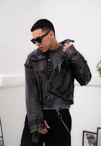 Black denim jacket with frayed edges and metal buttons, layered over a grey ribbed jumper. Accessories include sunglasses and a chain.