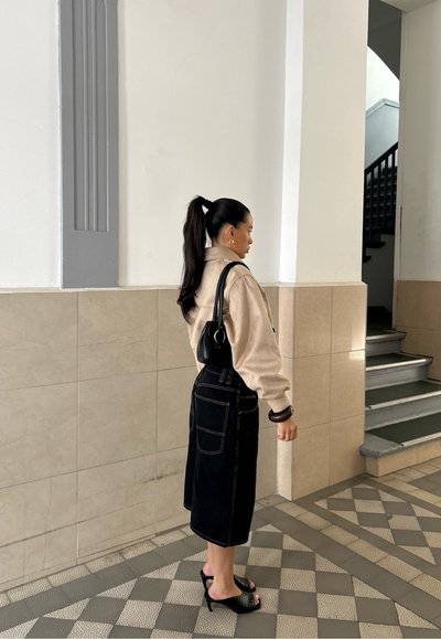 Sudadera de forro polar beige, falda de mezclilla negra con bolsillos, bolso negro y zuecos de tacón, de pie sobre un azulejo decorado en una escalera.