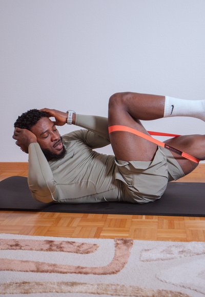 Hombre con una camisa de manga larga oliva clara y pantalones cortos grises hace ejercicio en una alfombra negra, utilizando una banda de resistencia naranja alrededor de las piernas.