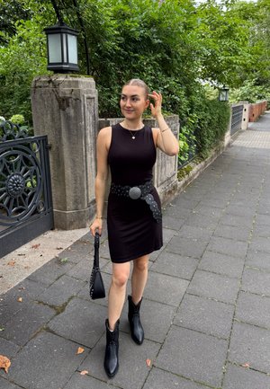 Joven mujer con un vestido negro sin mangas, cinturón ancho y botas negras, sosteniendo un bolso, de pie en una acera arbolada cerca de una puerta de piedra.