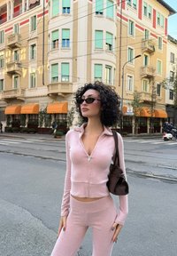 Różowy krótki sweter na zamek oraz dopasowane legginsy z wysokim stanem wykonane z miękkiego materiału, zestawione z ciemnobrązową torebką i czarnymi okularami przeciwsłonecznymi.