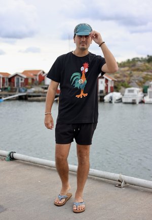 Camiseta negra con un gráfico colorido de gallo, pantalones cortos negros y una gorra azul de patrones. Las sandalias de diseño sencillo completan el look.