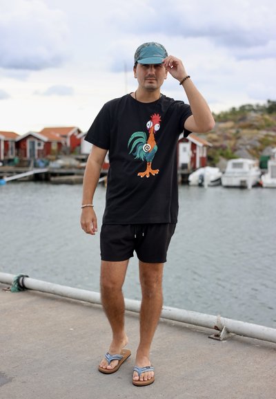 Camiseta negra con un gráfico colorido de gallo, pantalones cortos negros y una gorra azul de patrones. Las sandalias de diseño sencillo completan el look.