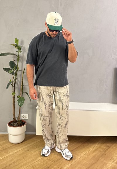 Conjunto casual que incluye una camiseta de gran tamaño en gris oscuro, pantalones beige con un patrón abstracto y zapatillas blancas. Los accesorios incluyen una gorra en beige y verde.