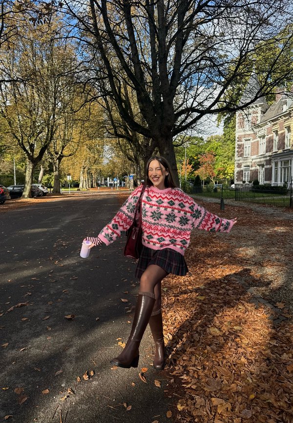 Une femme souriante pose les bras écartés dans une rue automnale couverte de feuilles. Elle porte un pull rose à motifs, une jupe à carreaux et de hautes bottes brunes.