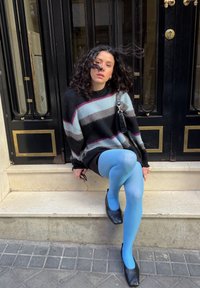 Άτομο με σγουρά μαλλιά, με ριγέ πουλόβερ, μπλε κολάν και μαύρα παπούτσια, καθισμένο σε εξωτερικά σκαλοπάτια μπροστά από μαύρη πόρτα με χρυσές λεπτομέρειες.