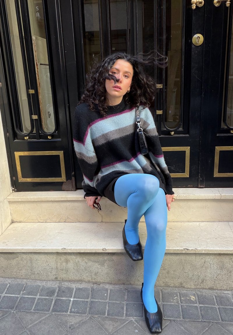 Krøllet person iført stribet sweater, blå leggings og sorte sko, siddende på udendørs trapper foran en sort dør med gulddetaljer.