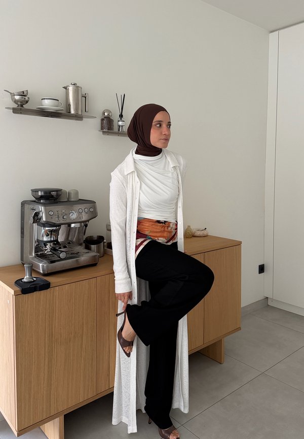 Une femme portant un hijab marron et un long cardigan blanc pose à côté d’une machine à café sur un plan de travail de cuisine en bois.