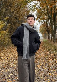 Juoda bombonešio striukė su pilku šaliku, kreminės kelnės, stovintis ant lapais nukloto tako tarp rudens medžių ir rūku apgaubtos fono.