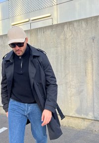 Μαύρο παλτό πάνω από μαύρο τζέρσεϊ, σε συνδυασμό με μπλε τζιν. Φοράει μπεζ καπέλο και γυαλιά ηλίου. Στον φόντο μια τσιμεντένια τοιχοποιία.