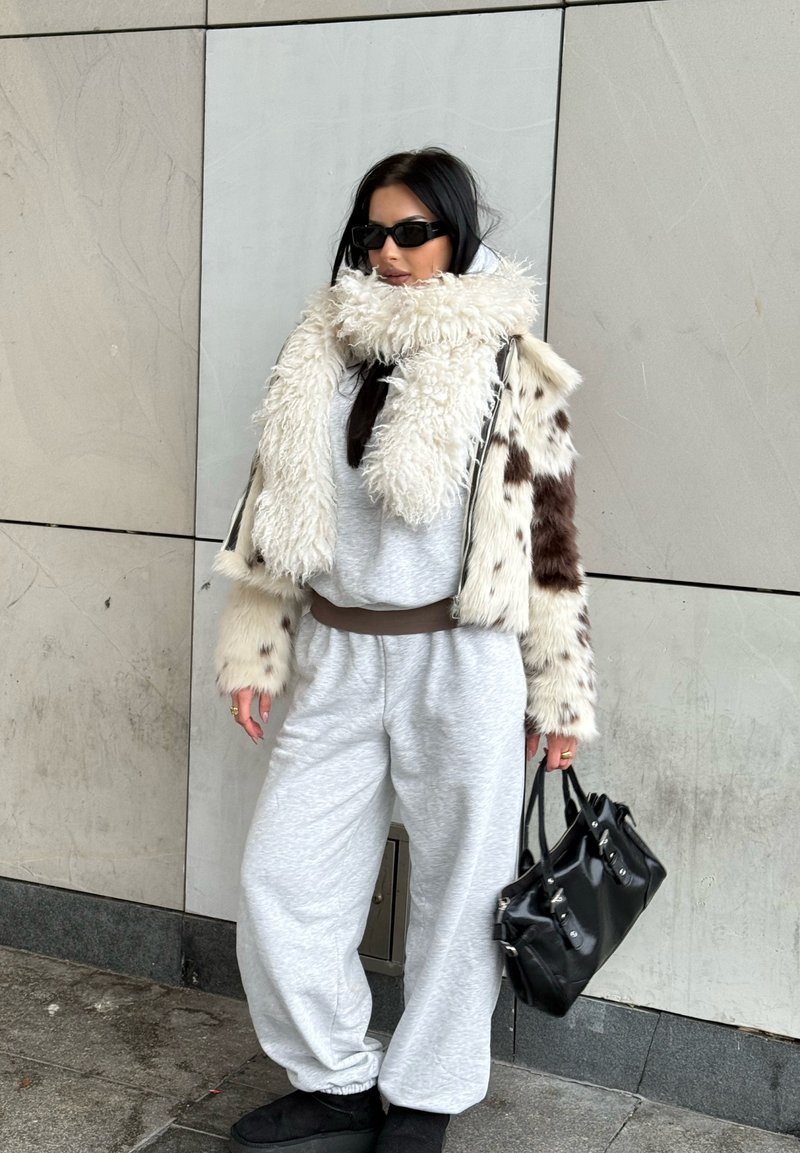 Personne porte une combinaison grise à taille cintrée, une veste en fourrure blanche et brune duveteuse, et des lunettes de soleil noires oversize, tenant un sac à main noir.