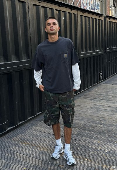 Camiseta de manga corta gris oscuro con un bolsillo en el pecho, superpuesta a una camisa de manga larga blanca. Pantalones cortos de camuflaje y zapatillas grises.