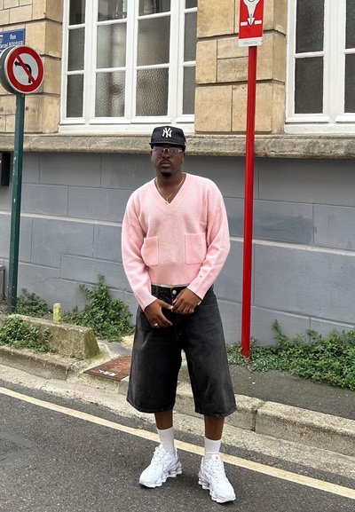 Suéter de punto rosa con escote en V y dos bolsillos frontales, combinado con pantalones cortos de mezclilla negros de gran tamaño y zapatillas deportivas blancas. Entorno urbano.