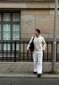 Ljust grått stickat tröja med V-ringning, vita byxor och en mörk axelväska, går på en kullerstensgata nära en stenvägg.