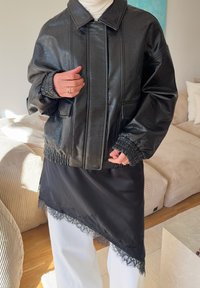 Veste oversize en simili cuir noir à finition texturée, avec poignets élastiques et deux poches avant, portée sur une jupe noire ornée de dentelle et un pantalon blanc.