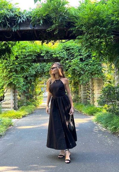 Vestido maxi negro de tipo halter con cintura fruncida y textura suave. La modelo lleva gafas de sol negras y sostiene un pequeño bolso, rodeada de vegetación.