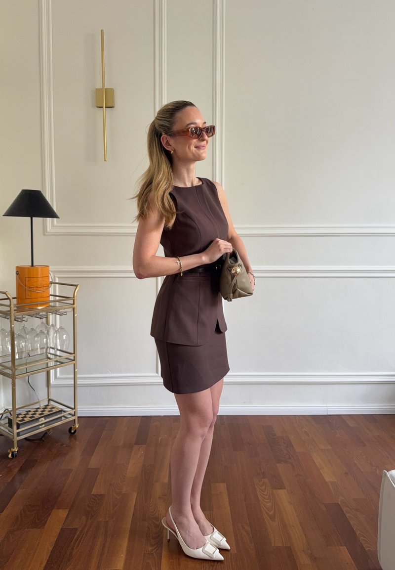 Nő ujjatlan barna ruhában és fehér magassarkúban, bézs kézitáskát tartva, egy fa padlószobában áll, ahol aranyszínű díszítésű italkocsi található.