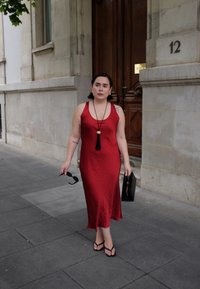 Donna che cammina su un pavimento di pietra vicino a un edificio, indossando un vestito rosso senza maniche, infradito neri, mentre tiene occhiali da sole e una borsa nera.