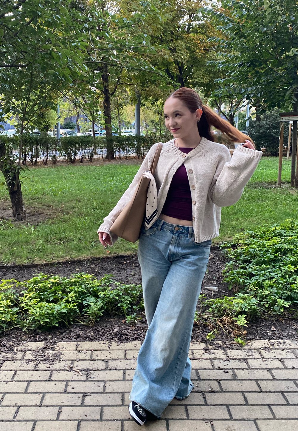 Svetlo béžový cardigan s gombíkmi, tmavofialový top, voľné svetlo modré džínsy, čierne tenisky, nesúci béžovú tašku s patterned šálom.