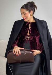 Haut en velours bordeaux avec des liens au niveau du cou, porté sous un manteau noir ; tenant un sac à main en cuir marron au design arrondi.