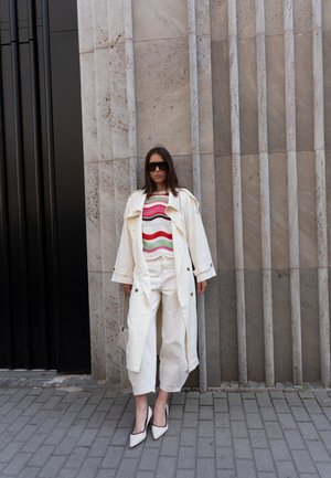 Mujer con abrigo trench oversize color crema, suéter de rayas, pantalones crema de pierna ancha y tacones blancos, de pie contra una pared de piedra con ranuras verticales.