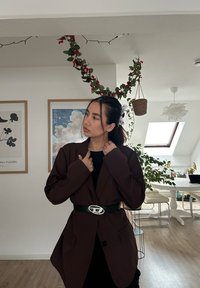 Blazer oversize marron foncé en tissu texturé, avec un unique bouton et des poches à l'avant, associé à une ceinture noire et un haut noir.