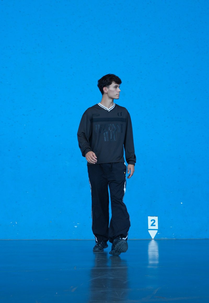Jeune homme portant un survêtement noir marchant à l'intérieur sur un sol réfléchissant devant un mur bleu vif avec un marqueur blanc numéro deux.