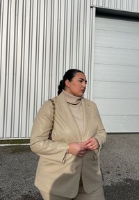 Μπεζ δερμάτινη σακάκι πάνω από ένα ανοιχτόχρωμο τζάκετ, συνδυασμένο με ταιριαστά παντελόνια. Αναδεικνύεται από μια τσάντα με αλυσίδα και ελάχιστα κοσμήματα.