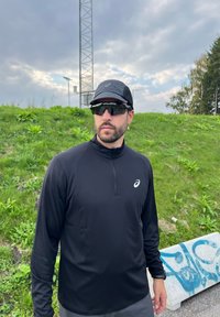 Melna sporta krekls ar garām piedurknēm un rāvējslēdzēja apkakli, melna cepure un saulesbrilles. Zāles fons ar betona barjeru un grafiti detaļu.