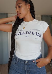 Hvit tettsittende t-skjorte med "MALDIVES" trykket i marineblått, kort ermer, kombinert med denimshorts. Modellen har langt flettehår og minimal smykker.