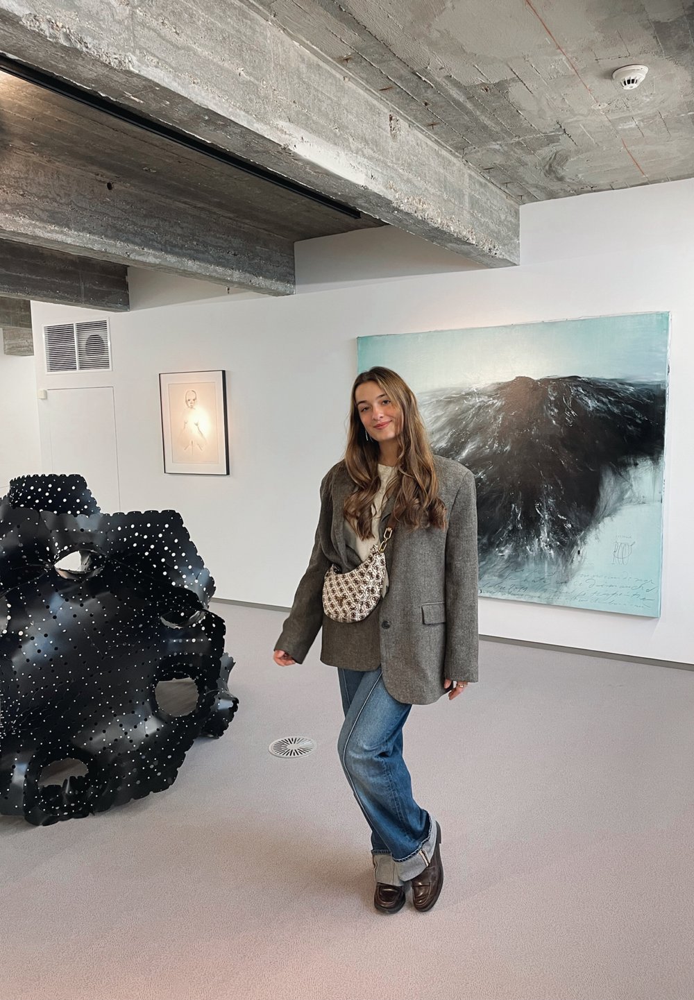 Grey oversized blazer, beige jumper, blue jeans, and brown shoes. Woven cream bag. Black abstract sculpture with holes in the background.