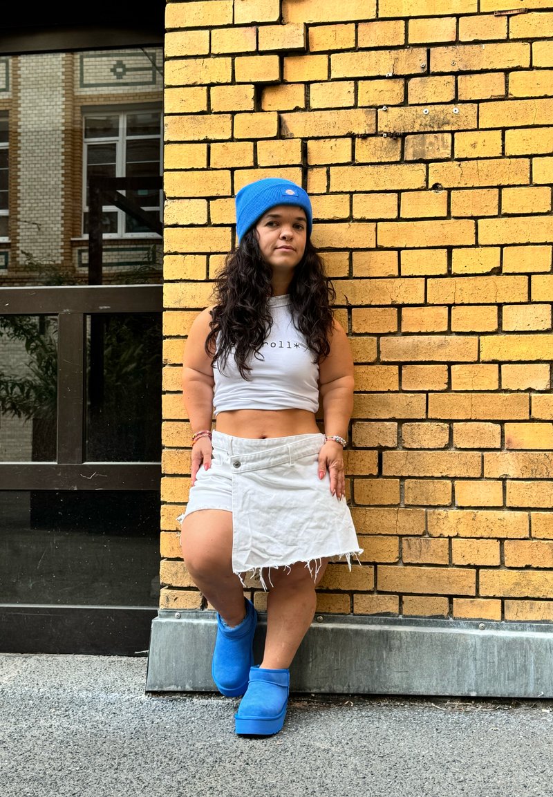 Μπλε σκουφάκι, λευκή κοντή μπλούζα, λευκή σχισμένη φούστα και μπλε πλατφόρμες, μπροστά σε υφασμάτινο κίτρινο τοίχο από τούβλα.