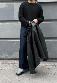 Άτομο με μαύρο πουλόβερ με υφή και φαρδιές μπλε τζιν, κρατώντας μαύρο δερμάτινο μπουφάν, στέκεται σε πλακόστρωτο πεζοδρόμιο δίπλα σε γκρίζο τοίχο.