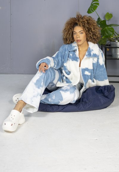 Chaqueta de forro polar con un patrón de nubes en azul y blanco, combinada con pantalones de tie-dye azul. Zuecos blancos y un sillón de frijol oscuro en un entorno minimalista.