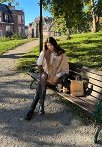 Ung kvinna i beige kappa och svarta stövlar sitter på en parkbänk med kaffemuggar och papperspåse, solig höstdag i stadsparken.