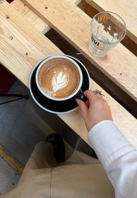 Sort kaffekopp med skål med lattekunst, plassert på en treflate. Ved siden av står et klart glass med logodesign.