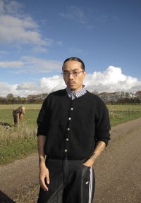 Černý chlupatý kardigan přes kostkovanou košili, v kombinaci s černými koženými kalhotami a logem Adidas. Přírodní pozadí s koněm v blízkosti.