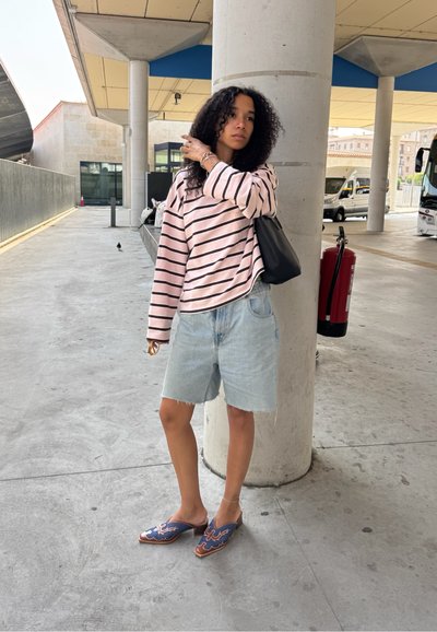 Top de manga larga a rayas en rosa claro y negro, pantalones cortos de mezclilla azul claro, zapatos azulones sin cordones con un patrón decorativo, bolso negro, de pie al aire libre.