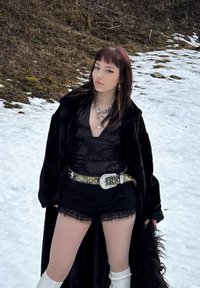 Nero cappotto in pelliccia sintetica, top nero con paillettes, pantaloni corti con pizzo, cintura decorata in argento, collane a strati, calze bianche fino al ginocchio, sfondo innevato.