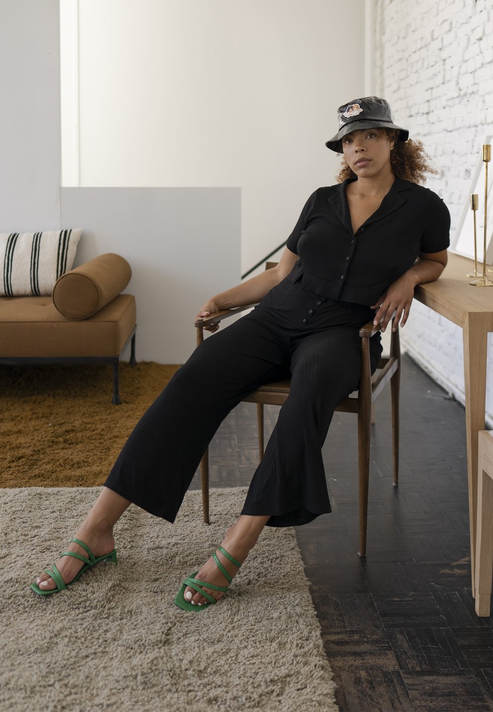 Zwarte geribde cropped blouse met knopen, bijpassende wijde broek en groene sandalen met bandjes. Lichte achtergrond, houten stoel en tapijt.