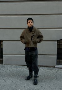 Geacă kaki scurtă cu guler înalt, purtată peste o cămașă închisă la culoare, asortată cu blugi negri largi și cizme din piele neagră, pe o trotuar gri.