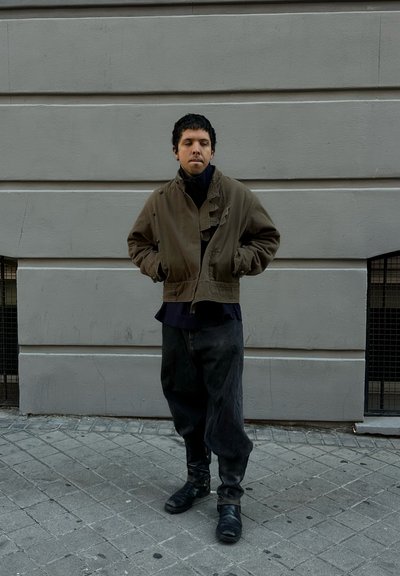 Chaqueta corta color caqui con cuello alto, usada sobre una camisa oscura, combinada con vaqueros negros holgados y botas de cuero negras en una acera gris.